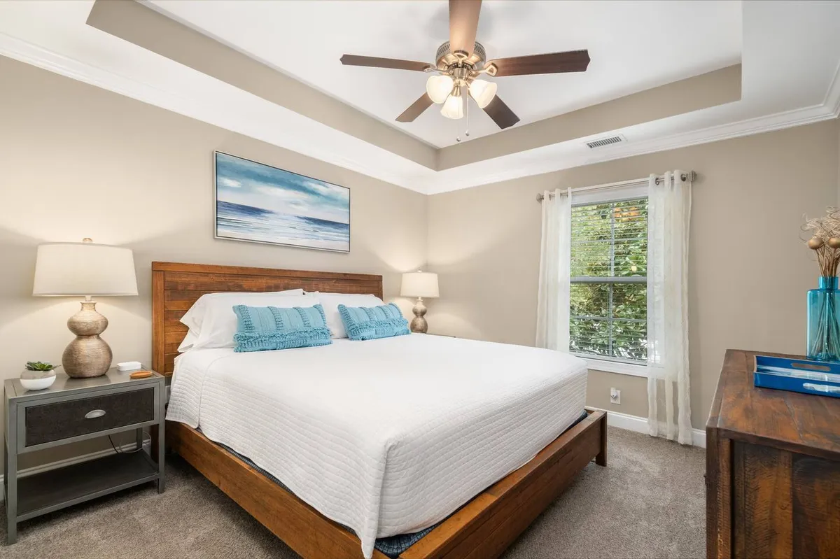 A cozy bedroom featuring a neatly made bed with blue cushions, a ceiling fan above, calming artwork positioned above the headboard, and a nightstand with a soft-glowing lamp. The room is bathed in natural light, creating a restful and inviting atmosphere ideal for relaxation.