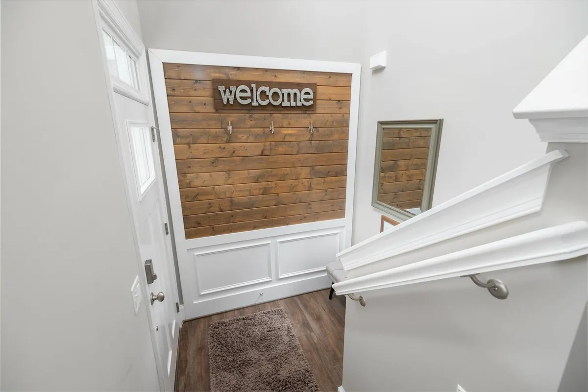 This image captures a warm, welcoming interior foyer with a wooden wall, a prominent welcome sign, coat hooks, and a mirror. The scene evokes the first impression guests receive upon entering, radiating coziness and hospitality.