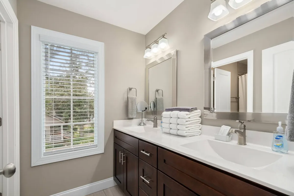 A modern bathroom featuring a double sink vanity with a large mirror, neatly organized towels, and a window allowing in morning light. The space feels fresh and spacious, ideal for relaxation and self-care routines.