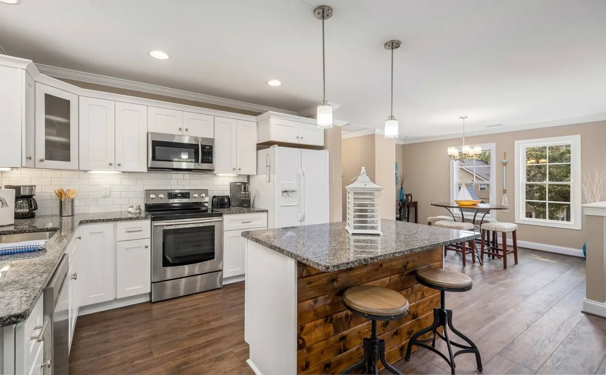This image captures an inviting interior with a granite kitchen island, stainless steel appliances, hanging light fixtures, and a dining area visible in the background. The open layout emphasizes both the kitchen and dining nook, with ample space for gathering, entertaining, and casual meals.
