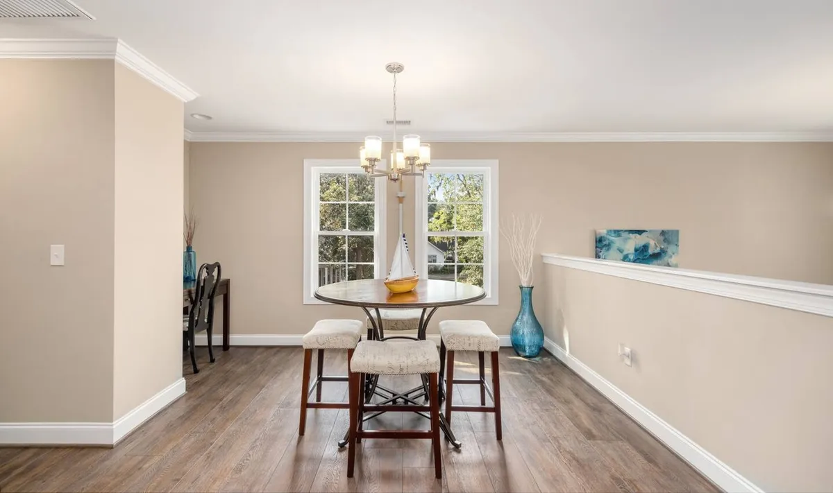 A welcoming dining nook with a round table, comfortable chairs, a chandelier, and large windows letting in natural light. Decorative touches like a vase and thoughtfully chosen decor create a cozy and intimate atmosphere, ideal for morning coffee or group meals.