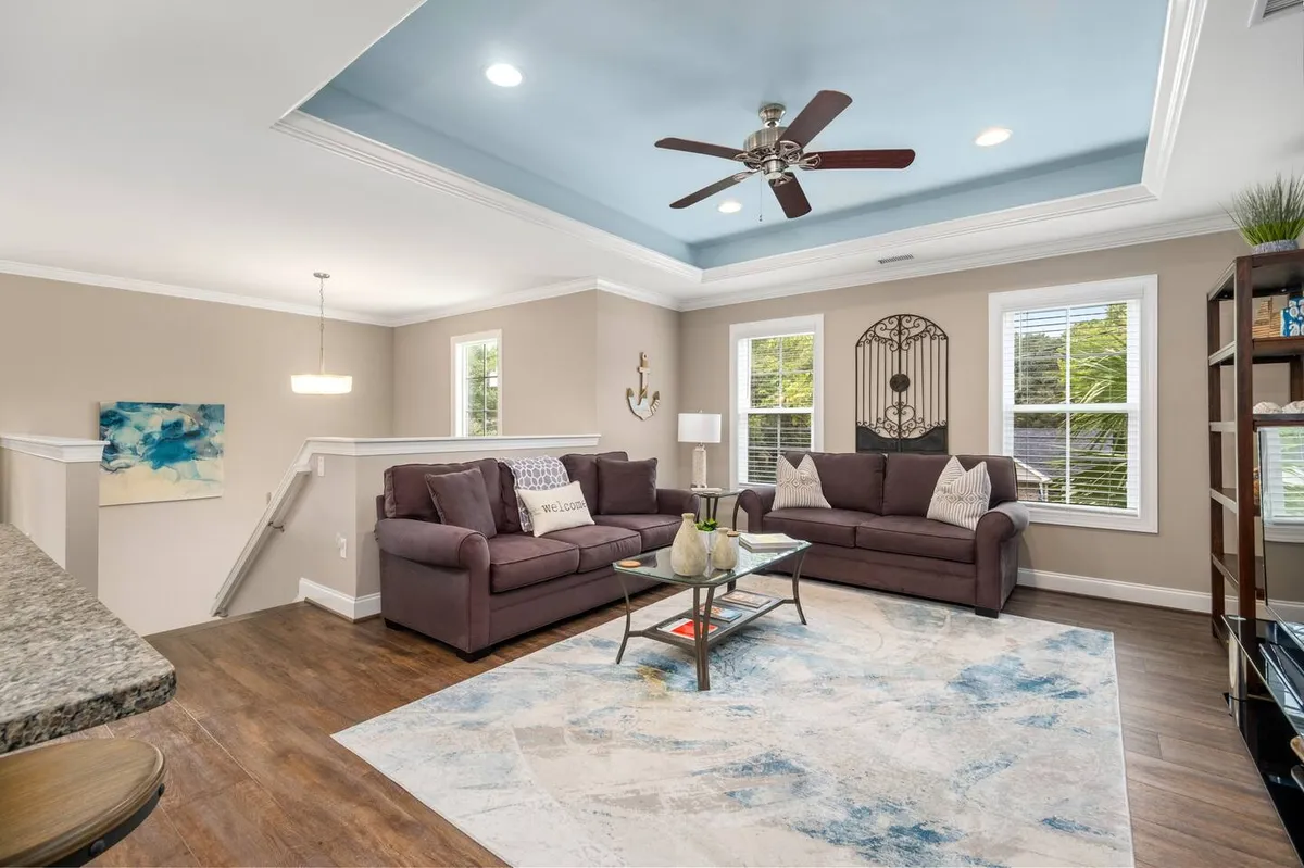 A bright, stylish living room featuring a comfortable sofa set, a chic coffee table atop a decorative rug, tasteful wall art, and a ceiling fan. The space radiates warmth and is designed for gatherings, relaxation, and casual entertaining.