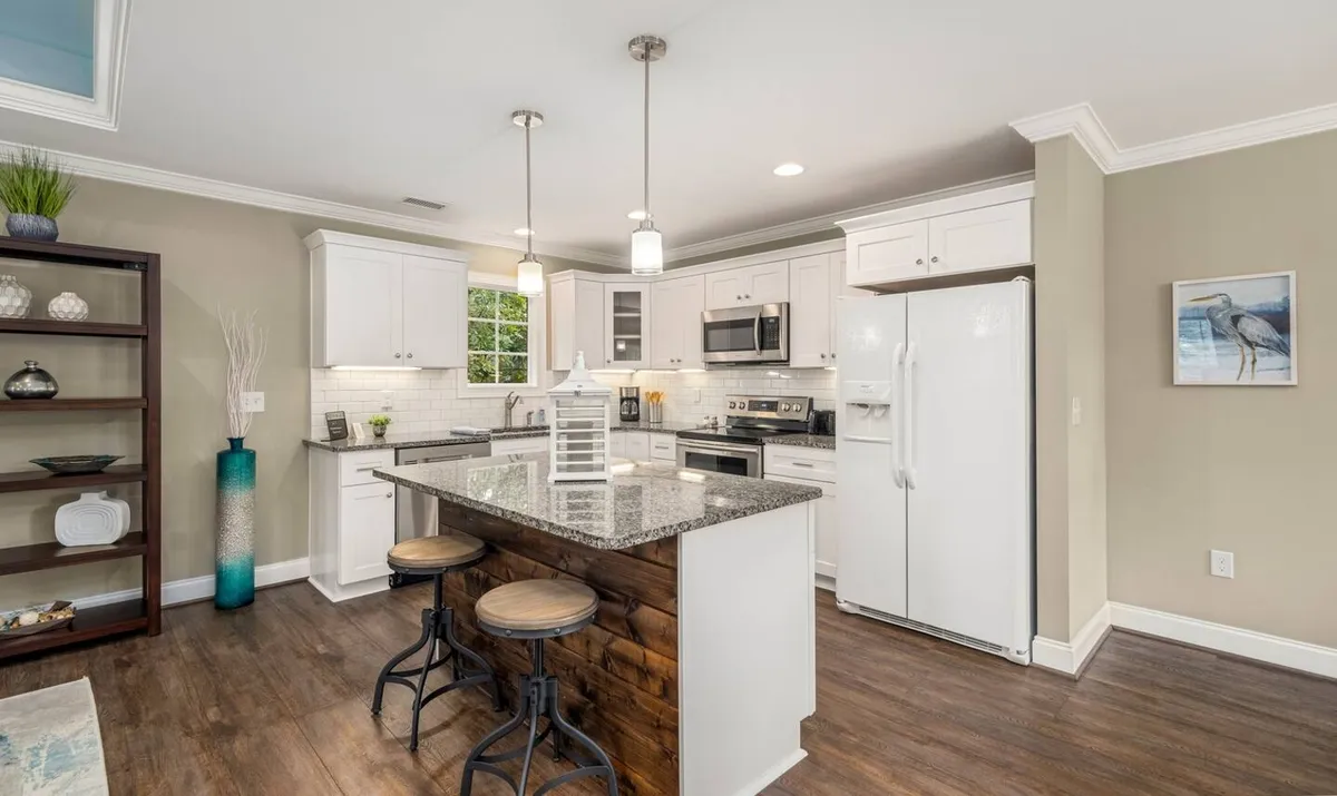 This image showcases a welcoming kitchen with a granite countertop, bar stools for casual seating, modern kitchen appliances, tastefully arranged shelf decor, and a window offering a view of lush greenery. The space balances functional, contemporary design with natural touches.